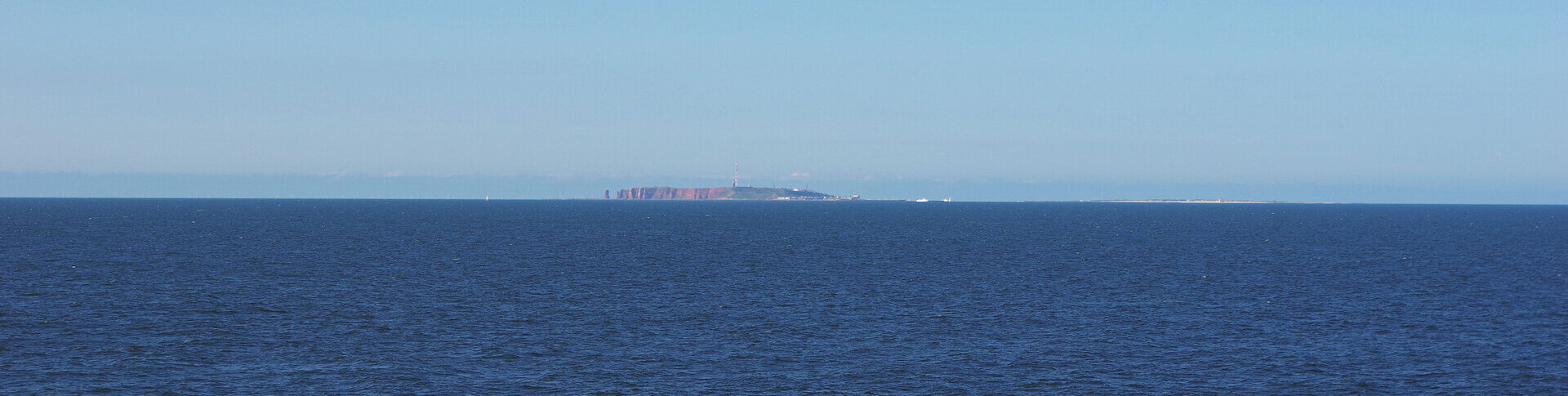 Weitwinkelaufnahme über das offene Meer mit Blick auf die Insel Helgoland am Horizont, mit der roten Steilküste und einem hohen Sendemast. Rechts von Helgoland ist die Düne zu sehen. 