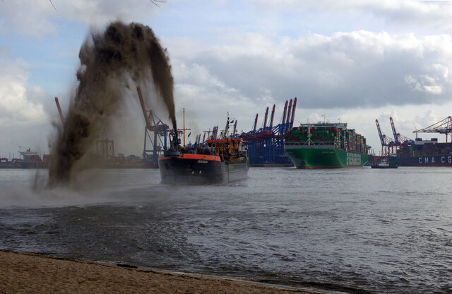 Ein Baggerschiff sprüht Sand und Wasser in hohem Bogen auf den Elbstrand, im  Hintergrund ein Containerschiff und Hafenterminals