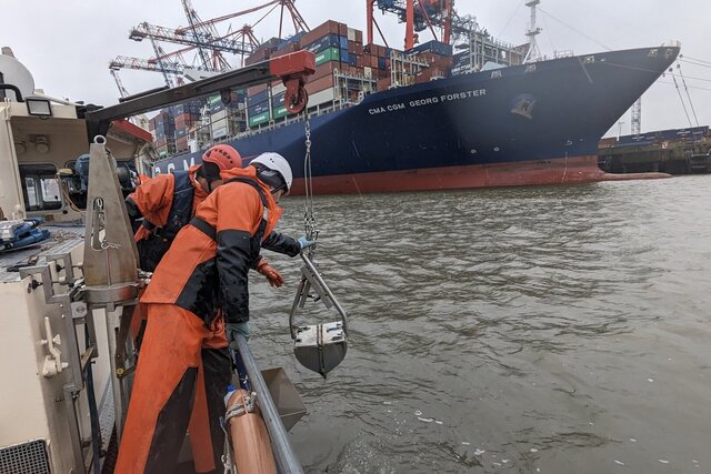 Zwei Männer in Ölzeug entnehmen eine Sedimentprobe aus einem Hafenbecken in Hamburg mit einem Greifer . Im Hintergrund ist ein Containerschiff und Containerbrücken