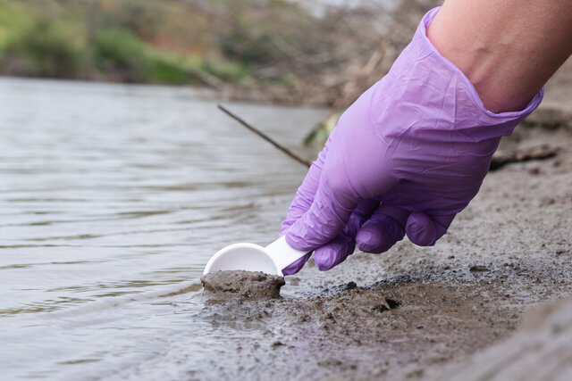 Eine Hand mit lila Handschuhen entnimmt mit einem kleinen Löffel eine Sedimentprobe aus einem Gewässer