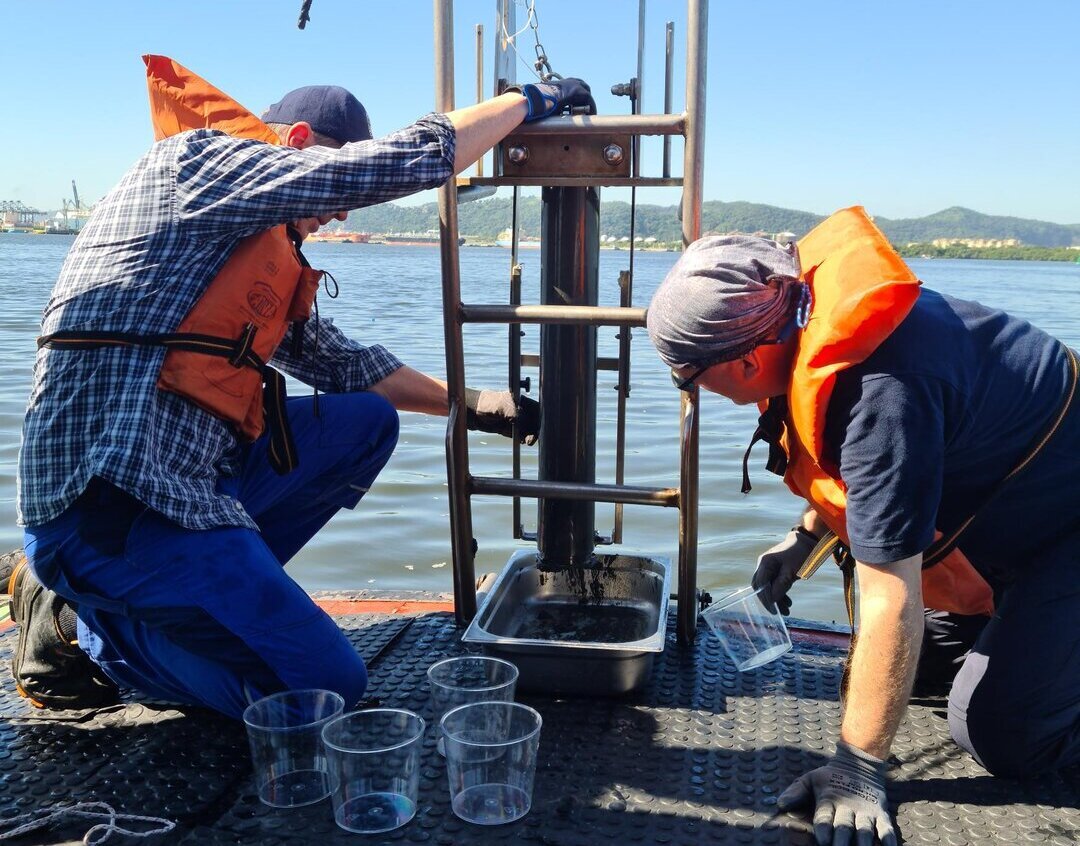 Zwei Wissenschaftler der HPA bedienen an Bord eines Schiffes ein kompliziertes Gerät zur Entnahme von Sedimentproben, im Hintergrund Hafen von Santos in Brasilien