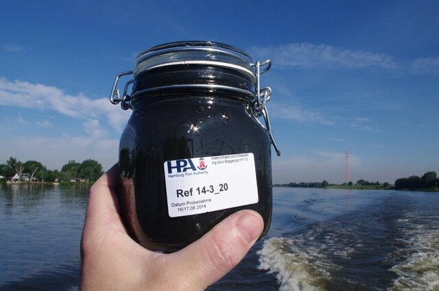 Eine Hand hält ein Probengefäß mit Schlick in die Kamera. Auf dem Glas ist ein Etikett der Hamburg Port Authority (HPA) mit der Aufschrift „Ref 14-3_20“ sowie dem Probedatum „16/17.08.2014“ zu sehen. Im Hintergrund ist die Elbe zu erkennen.