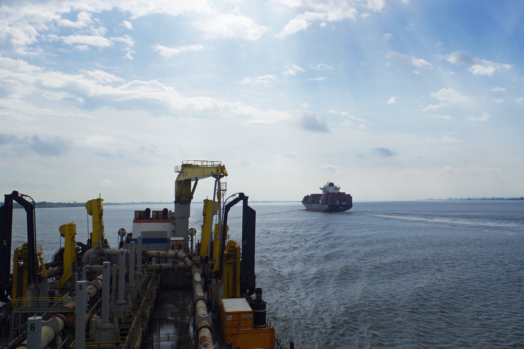Blick von der KAISHUU auf ein Containerschiff, das elbaufwärts in Richtung Hamburg fährt. Der Vordergrund zeigt technische Ausrüstung und Rohrleitungen auf dem Schiff. Der Himmel ist leicht bewölkt, die Sonne scheint durch die Wolken.