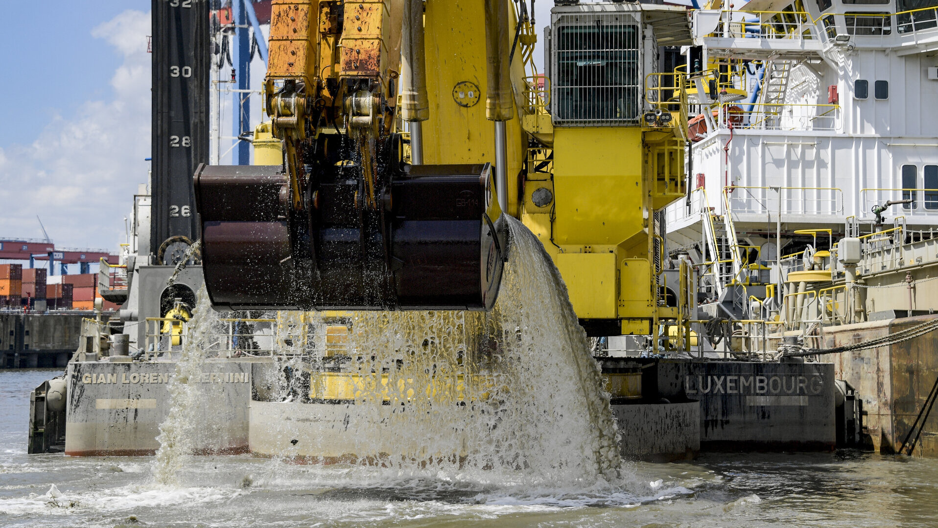 Eine große Baggerschaufel eines gelben Schwimmbaggers baggert Sediment aus der Elbe im Hamburger Hafen. Das Baggerschiff trägt den Namen „Gian Lorenzo Bernini“ und ist in Luxemburg registriert. Auf der linken Seite sind gestapelte Container zu sehen.
