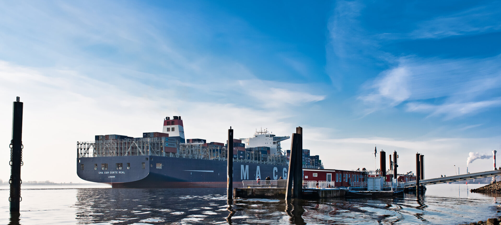 Ein großes blaues Containerschiff mit dem Namen CMA CGM Corte Real auf der Elbe. Im Vordergrund ist ein schwimmender Anleger mit einem Haus und Menschen. Im glatten Wasser stehen Pfähle , rechts ein Leuchtturm und ein Schornstein. Der Himmel ist blau mit leichter Bewölkung.