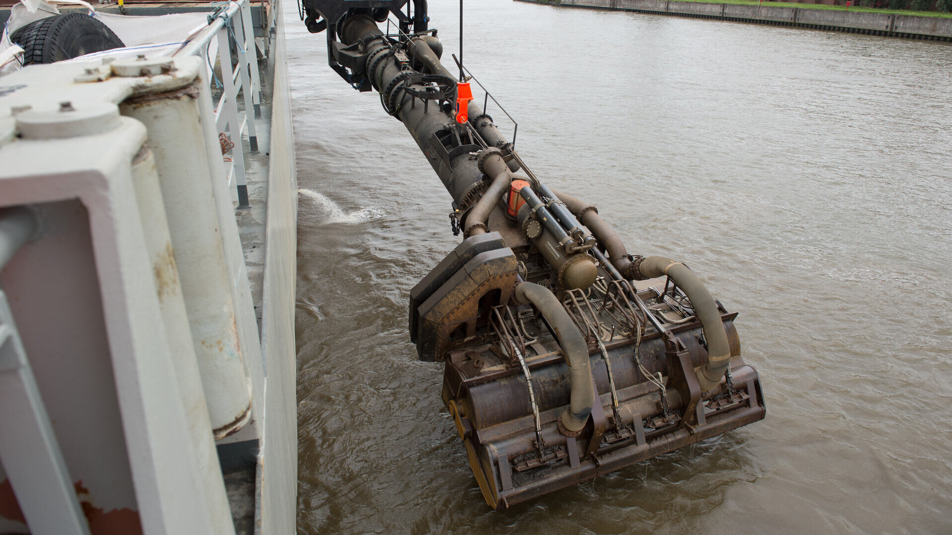 Nahaufnahme eines Hopperbaggers im Einsatz. Der Saugkopf, ein großes, industrielle Gerät ist ins Wasser abgesenkt und zeigt komplexe Schläuche, Metallstrukturen und Mechanismen für den Baggerbetrieb.