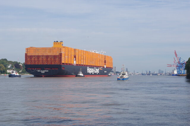 Ein großes Containerschiff mit dem Namen „Berlin Express“ fährt die Elbe hinauf in Richtung Hamburger Hafen, begleitet von mehreren Schleppern. Im Hintergrund sind Hafenkräne, die Skyline Hamburgs und die Elbphilharmonie zu sehen.
