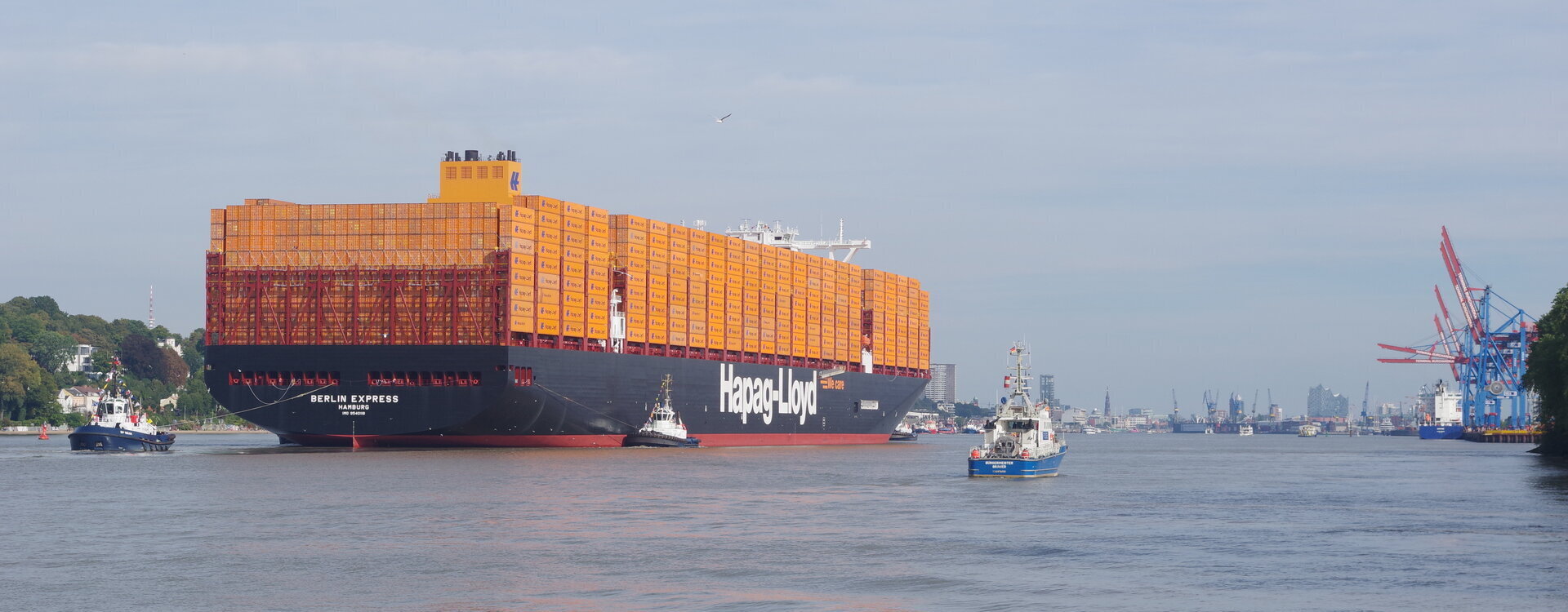 Ein großes Containerschiff mit dem Namen „Berlin Express“ fährt die Elbe hinauf in Richtung Hamburger Hafen, begleitet von mehreren Schleppern. Im Hintergrund sind Hafenkräne, die Skyline Hamburgs und die Elbphilharmonie zu sehen.