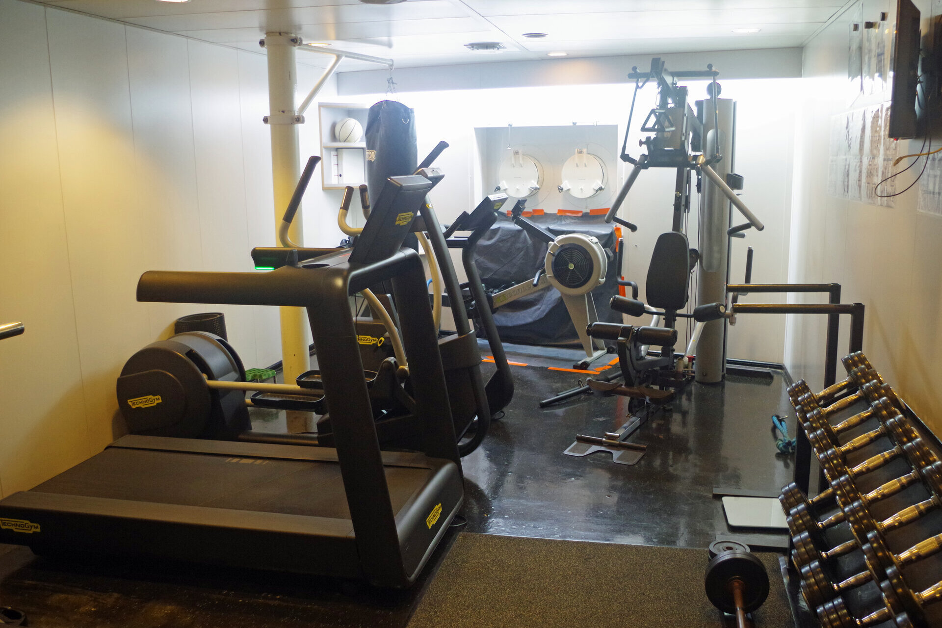 Ein kompakter Fitnessraum mit verschiedenen Trainingsgeräten, darunter ein Laufband, ein Crosstrainer, ein Rudergerät, eine Multifunktions-Kraftstation und eine Hantelbank mit Hantelset. Im Hintergrund sind ein Boxsack und zwei runde Bullaugen sichtbar.