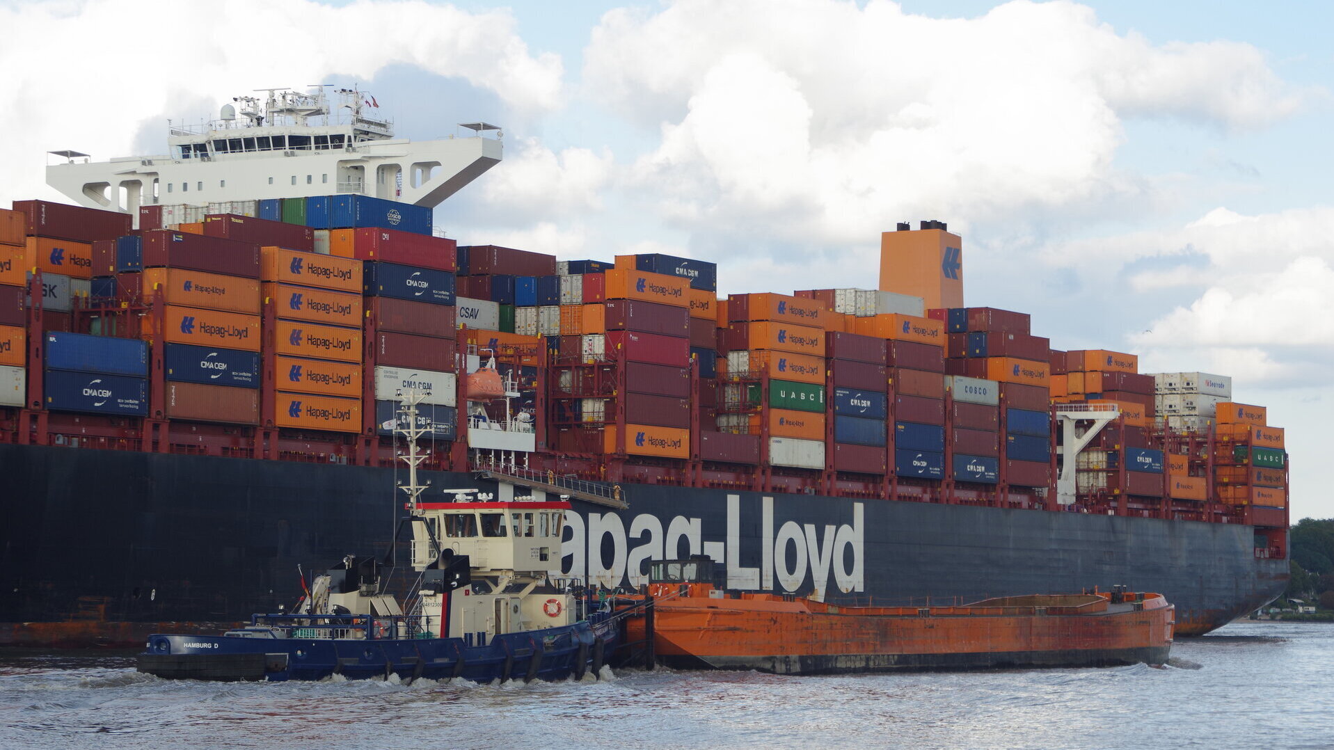 Ein großes Containerschiff mit vielen bunten Containern beladen im Hamburger Hafengebiet, im Vordergrund ein Schubverband für Baggergut.