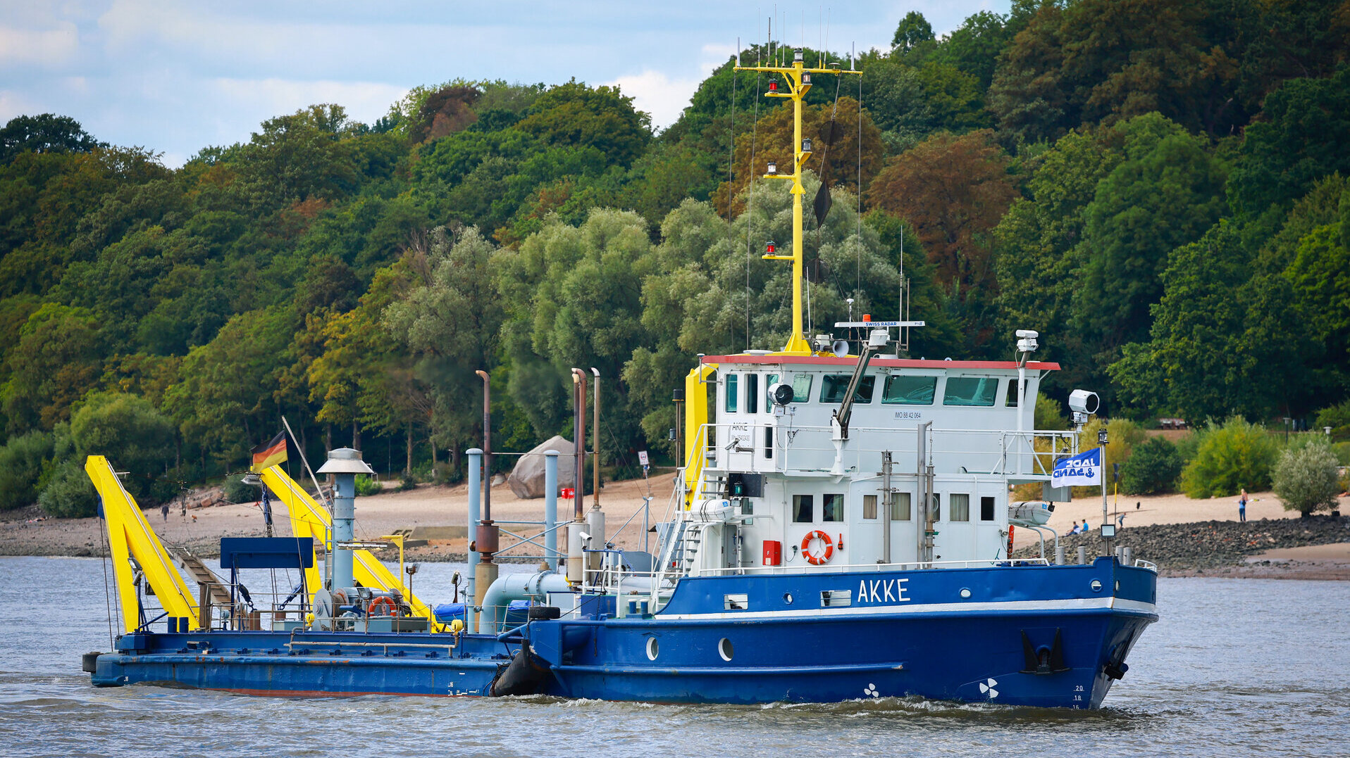 Das blaue Baggerschiff „AKKE“, ein Wasserinjektionsgerät, mit weißer Brücke und gelbem Mast fährt auf der Elbe. Im Hintergrund ist ein bewaldetes Ufer mit Sandstrand und Spaziergängern zu sehen.