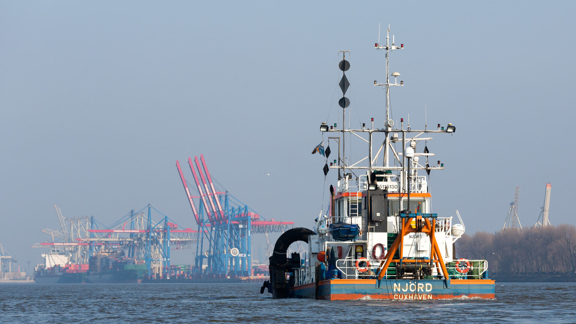 Ein Arbeitsschiff mit dem Namen „NJÖRD“ aus Cuxhaven fährt auf der Elbe im Hamburger Hafen. Es ist mit verschiedenen technischen Geräten und Signalmasten ausgestattet. Im Hintergrund sind große Hafenkräne und Containerschiffe zu sehen. 
