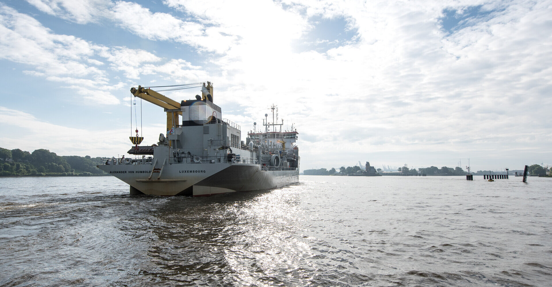 Ein großes Schiff, eine Hopperbagger mit dem Namen „Alexander von Humboldt“ und einem Kran, fährt auf der Elbe. Im Hintergrund ist eine Uferlandschaft und der Hamburger Hafen zu sehen. Der Himmel ist teils bewölkt und die Sonne spiegelt sich auf der Wasseroberfläche.