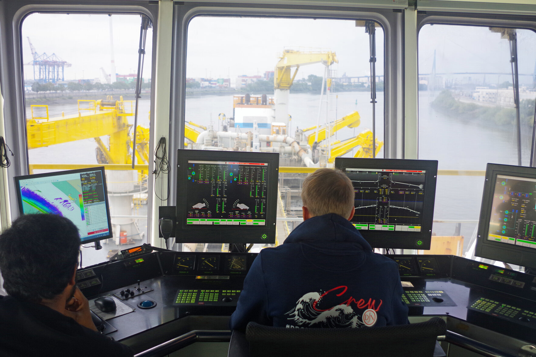 Zwei Personen sitzen an einem Steuerstand auf der Brücke des Baggerschiffs KAISHUU und blicken auf mehrere Monitore mit technischen Anzeigen. Durch die Fenster ist der Laderaum und das Deck des Baggerschiffs mit Rohren und gelben Kränen zu sehen, dahinter der Hamburger Hafen mit Containerbrücken und der Köhlbrandbrücke.