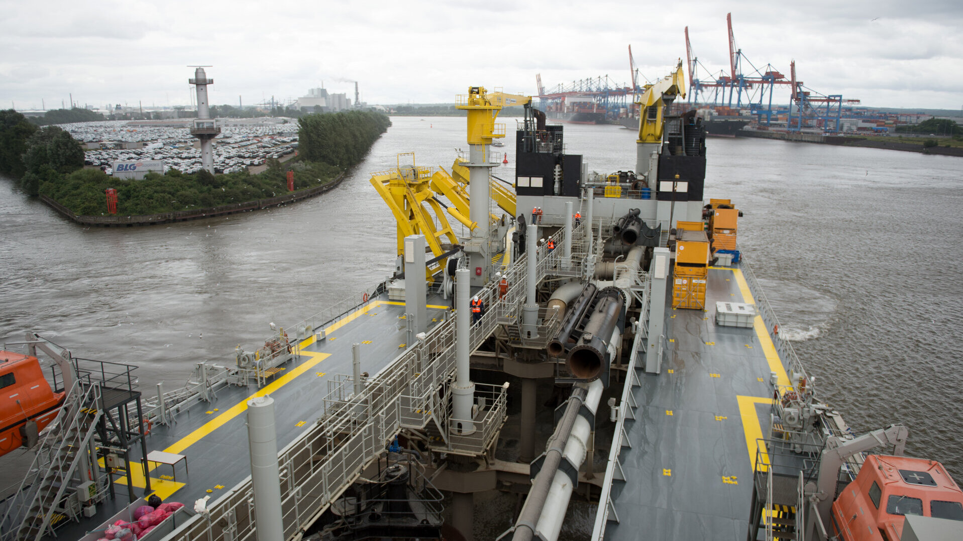 Deck eines Hopperbaggers mit großen Rohren, Kränen und Sicherheitsausrüstung. Mehrere Arbeiter in Sicherheitskleidung befinden sich auf dem Schiff, während im Hintergrund der Hamburger Hafen mit Containern und Kränen zu sehen ist.