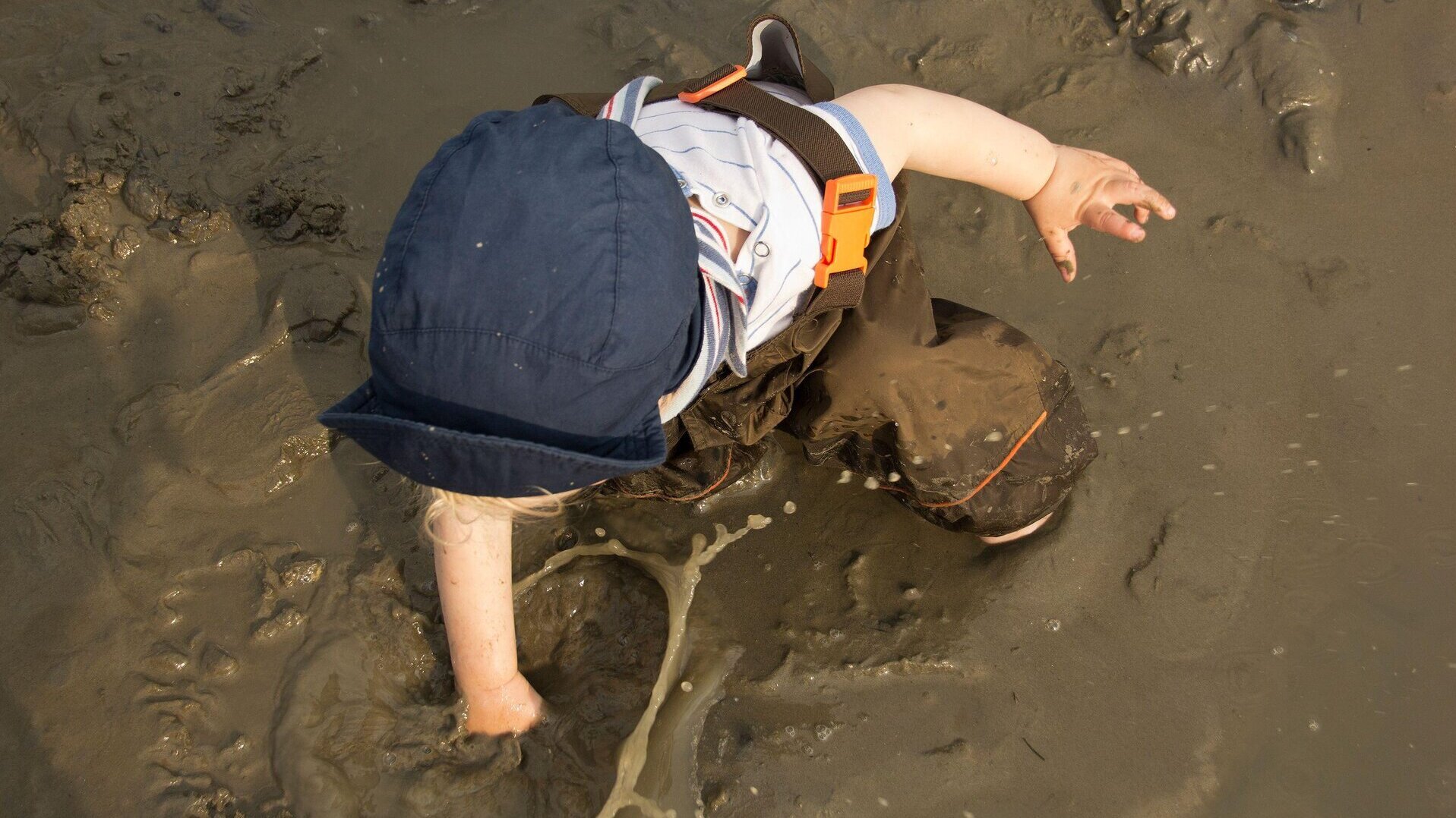 Ein Kind mit blauem Sonnenhut und Matschhose spielt im nassen Schlick, steckt eine Hand tief in den Boden und spritzt dabei Schlamm auf.