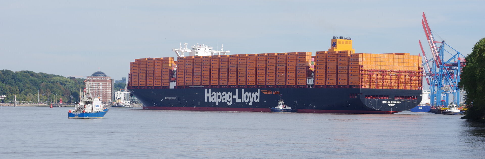 Das Containerschiff „Berlin Express“ wird mithilfe von Schleppern in der Elbe gedreht, um am Hamburger Hafen anzulegen. Im Hintergrund sind Containerkräne, Hafenanlagen und der Museumshafen Oevelgönne zu sehe.