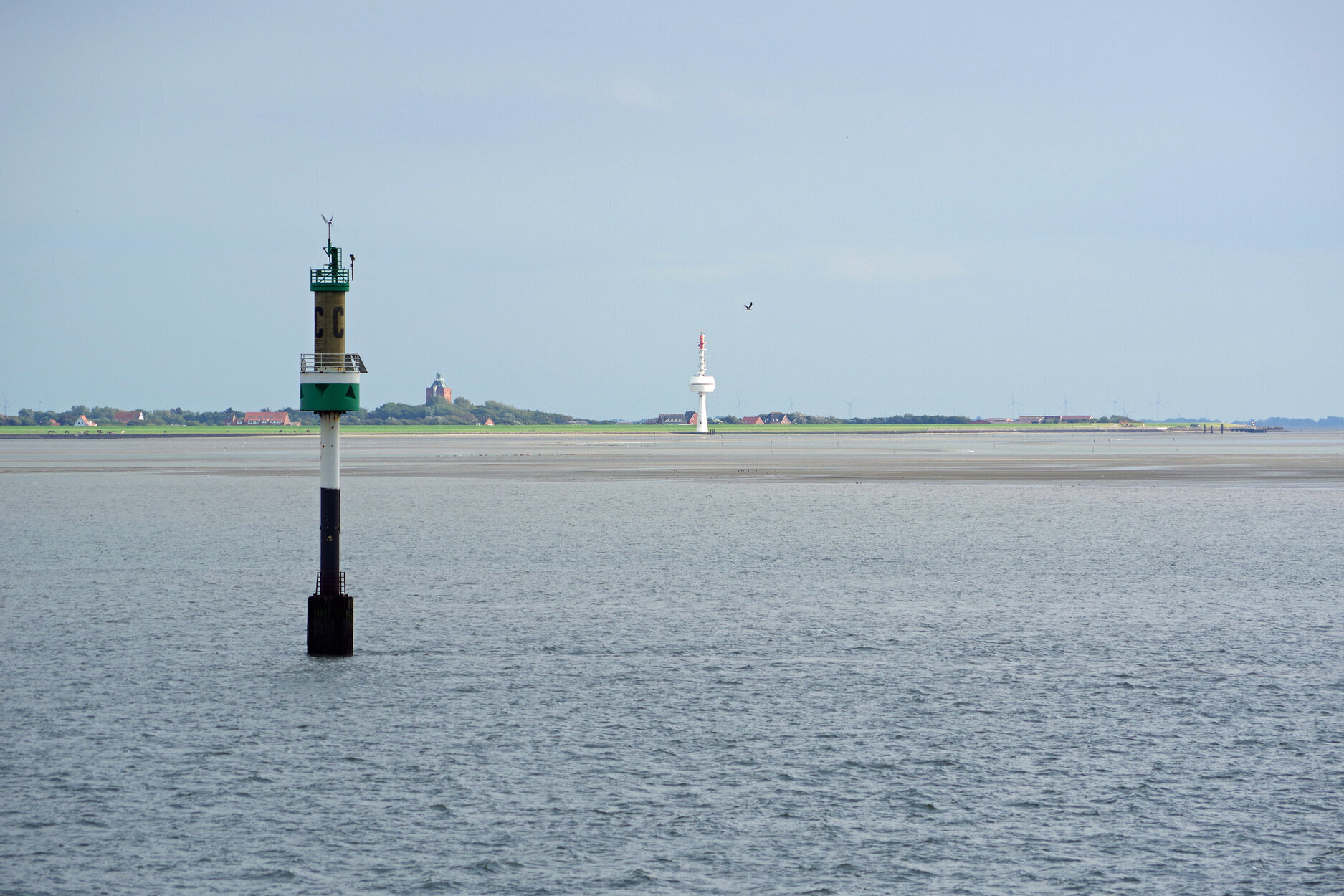 Seezeichen im Wasser mit grün-schwarzer Markierung im Vordergrund; im Hintergrund die Küstenlandschaft der Insel Neuwerk mit Gebäuden, einem rot-weißen Funkturm und dem historischen Leuchtturm, davor Watt und Rastvögel