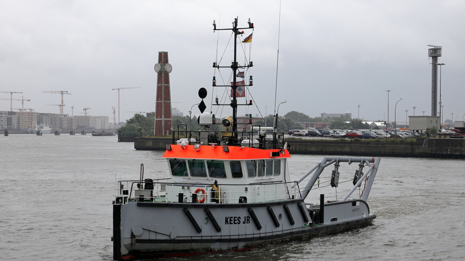 Ein Schlickpflug-Baggerschiff mit einer leuchtend orangenen Brücke und dem Namen „KEES JR“ fährt auf der Elbe im Hamburger Hafen. Es besitzt einen hohen Mast mit Signallichtern, Antennen und einer kleinen deutschen Flagge. Im Hintergrund sind ein markanter roter Uhrenturm, zahlreiche Baukräne und ein Parkplatz mit Autos zu sehen. Der Himmel ist grau und bewölkt.
