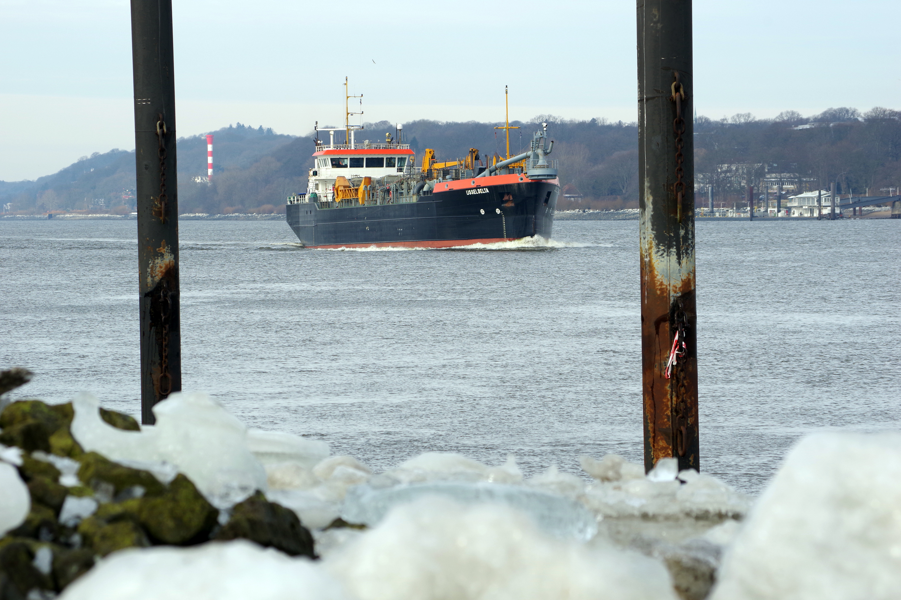 Der Hopperbagger Ijsseldelta fährt auf der Elbe, im Vordergrund sind Pfähle und Eis