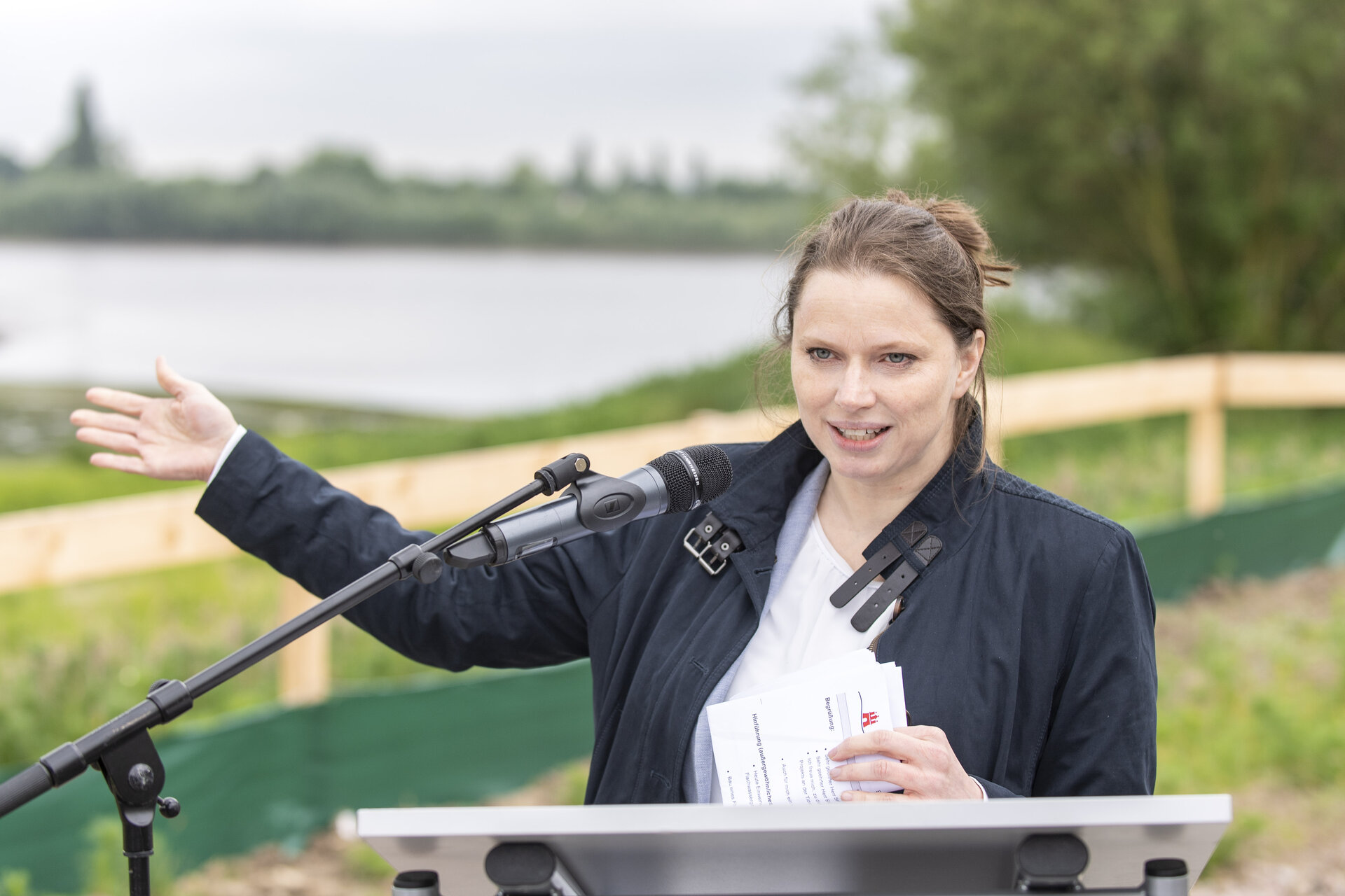 Die Hamburger Wirtschaftssenatorin Dr. Melanie Leonhard steht hinter einem Mikrophon und Rednerpult und zeigt auf eine Wasserfläche.