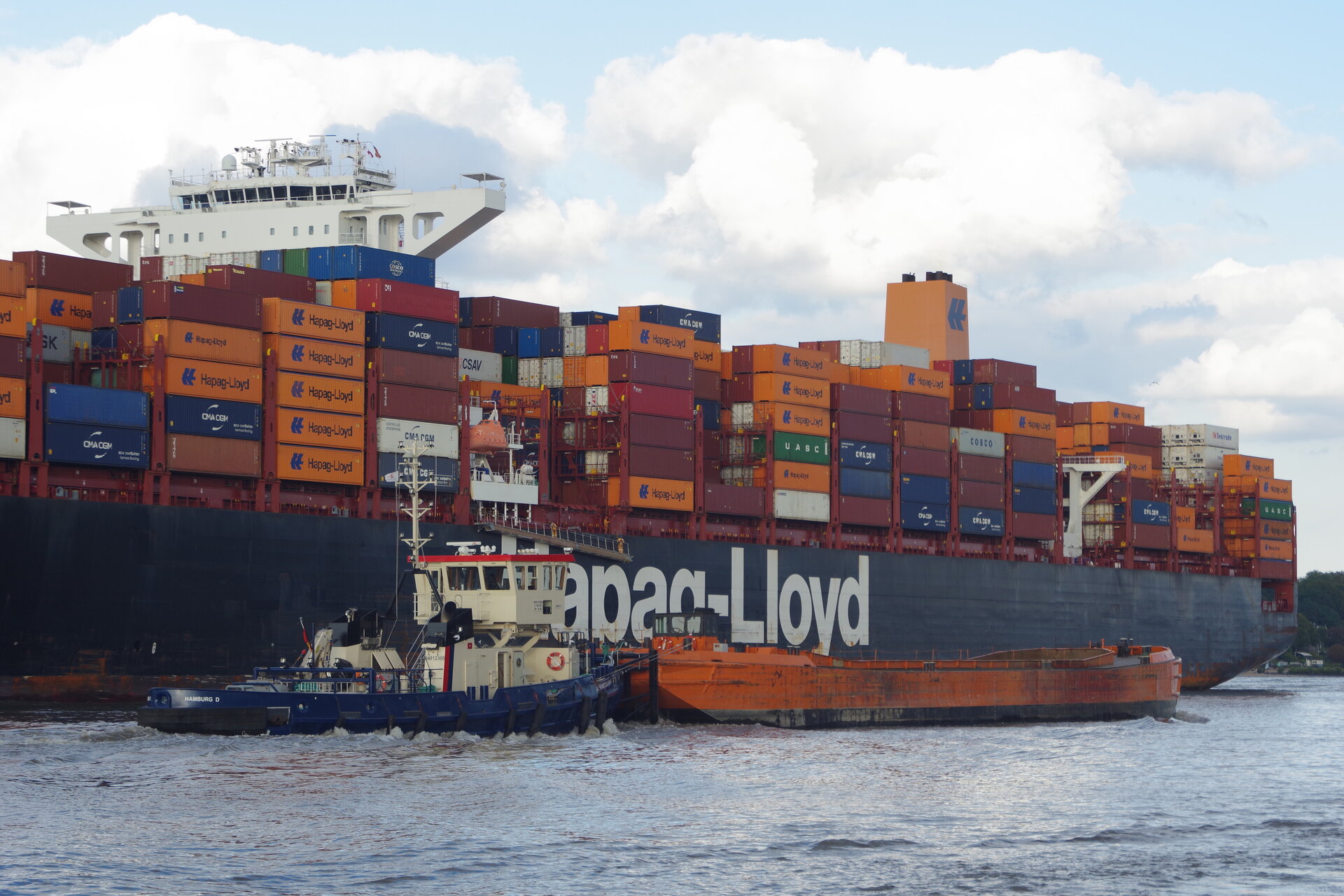Ein großes Containerschiff mit vielen bunten Containern beladen im Hamburger Hafengebiet, im Vordergrund ein Schubverband für Baggergut.