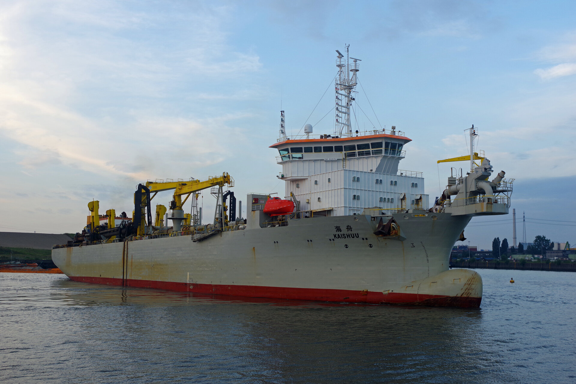 Das Baggerschiff KAISHUU liegt auf der Elbe, mit gut sichtbarer Brücke, markanter Aufbautenstruktur und mehreren gelben Kranarmen an Deck. Der Rumpf ist grau mit rotem Unterwasseranstrich, im Hintergrund ist ein Ufer mit Bäumen und Industrieanlagen des Hamburger Hafens zu sehen.