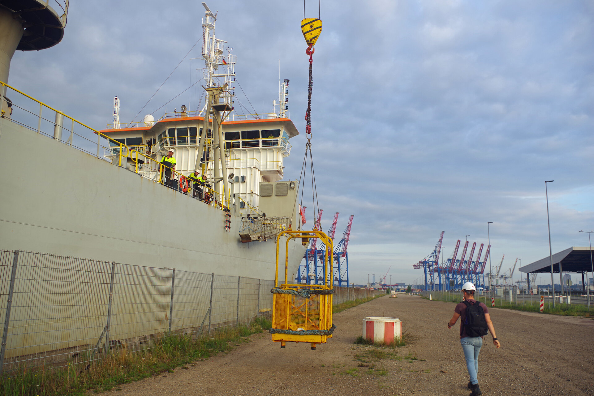 Ein gelber Ladekorb wird mithilfe eines Krans vom Baggerschiff KAISHUU auf den Kai gehoben. Mehrere Personen in Warnwesten stehen an der Reling des Schiffes, während eine weitere Person mit Schutzhelm und Rucksack auf dem Hafengelände entlangläuft. Im Hintergrund sind zahlreiche Containerkräne des Hamburg Hafens zu sehen.