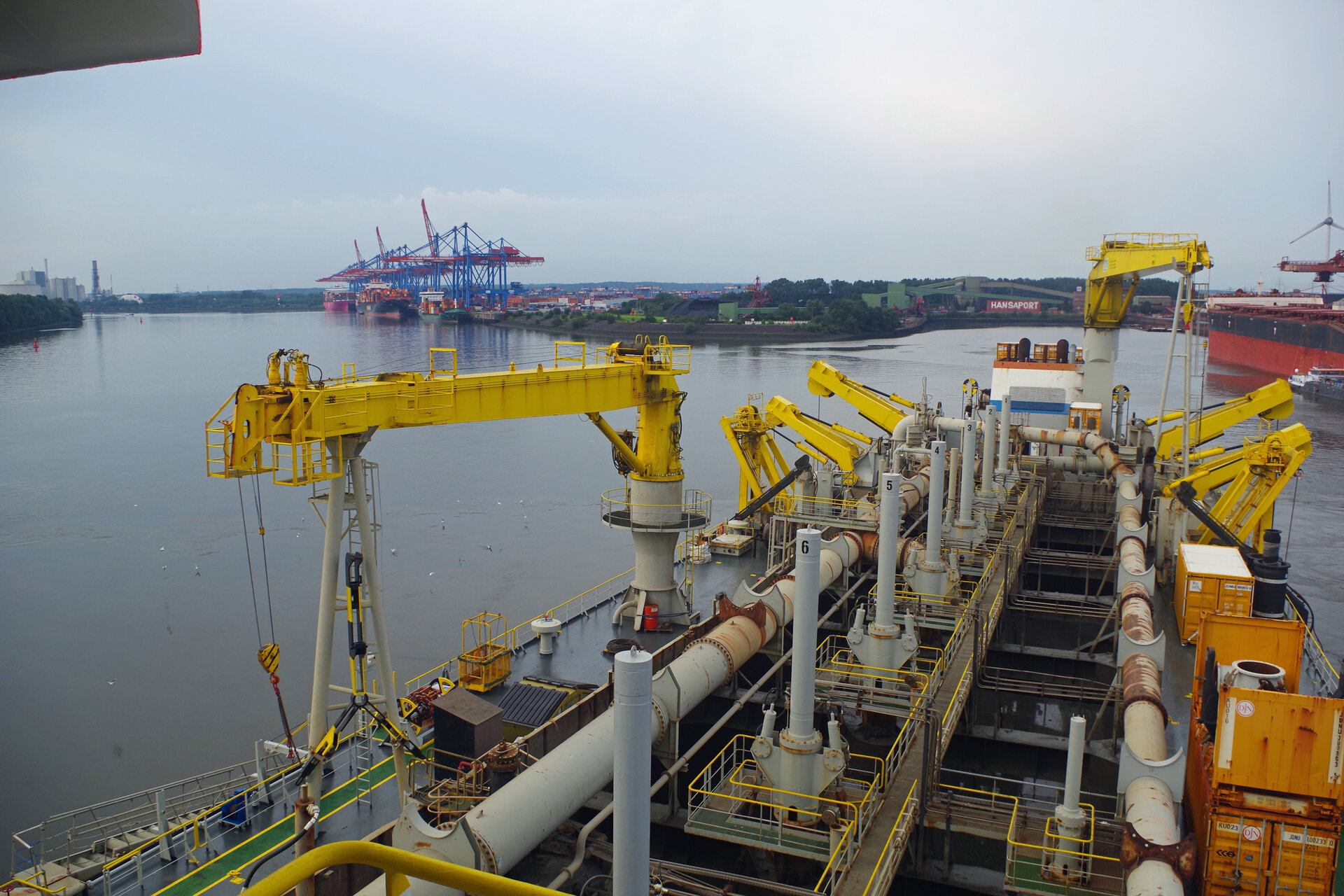 Blick vom Baggerschiff KAISHUU auf das Deck und den Laderaum des Baggerschiffs. Im Hintergrund die Süderelbe mit dem Containerterminal Altenwerder der HHLA und das Massengutterminal Hansaport im Sandauhafen. 
