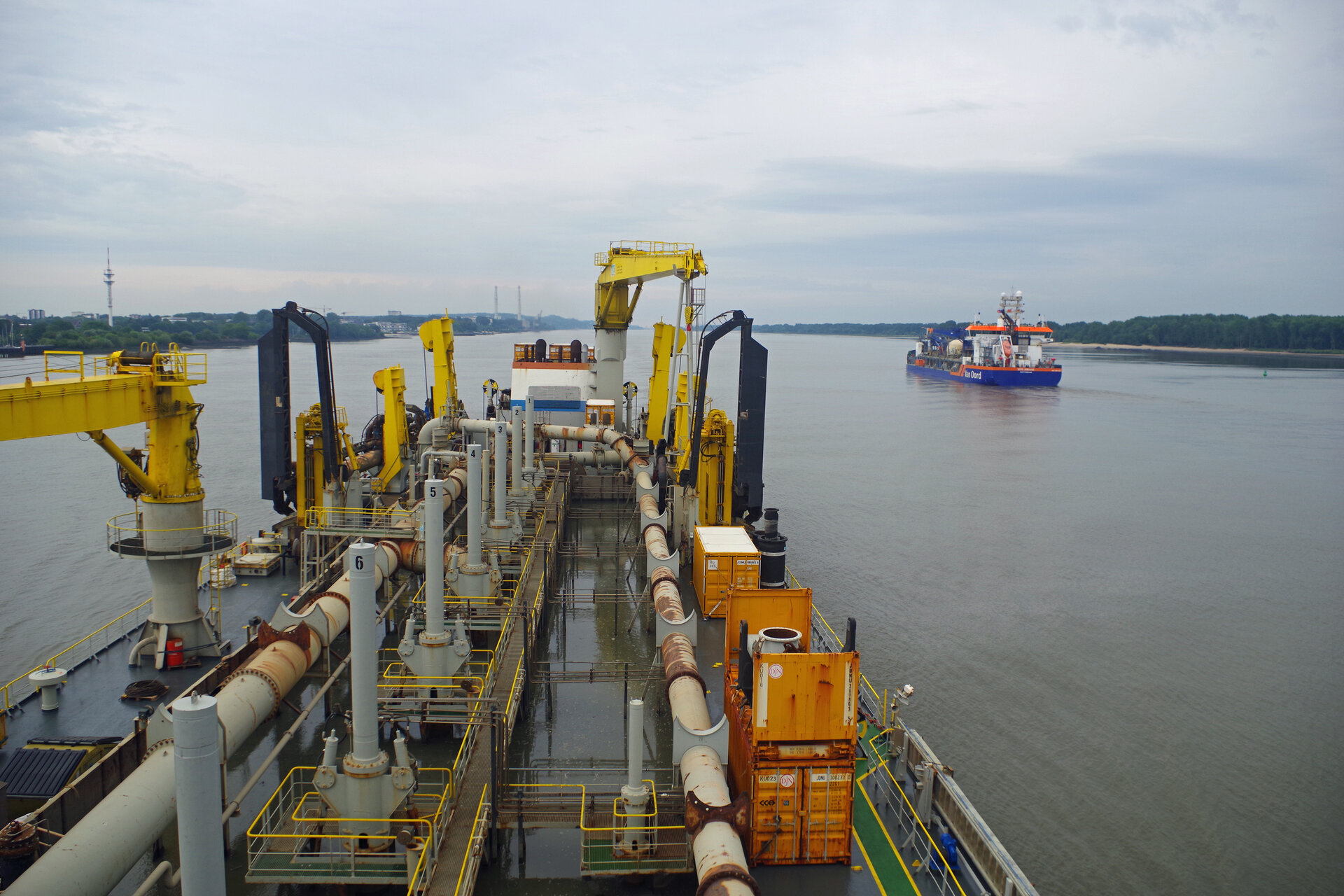 Blick vom Deck des Baggerschiffs KAISHUU mit zahlreichen Rohrleitungen, Maschinen und gelben Kränen auf ein weiteres Baggerschiffschiff auf der Elbe. Am Horizont ist eine grüne Uferlandschaft das Heizkraftwerk Wedel und ein Radarturm zu sehen. Der Himmel ist bewölkt.