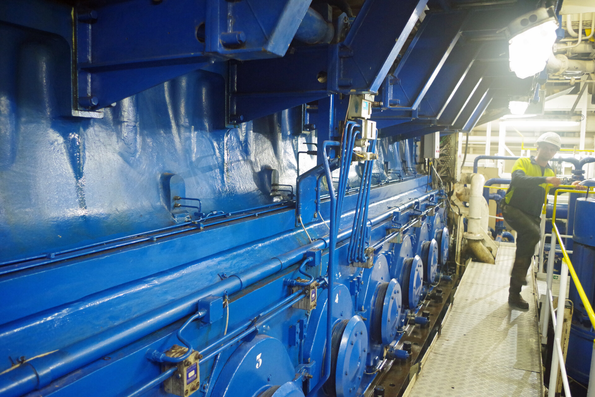 Großer blauer Schiffsdieselmotor im Maschinenraum der KAISHUU, flankiert von einer begehbaren Plattform. Ein Techniker mit Helm und Warnkleidung bewegt sich über den schmalen Gang entlang des Motors. 