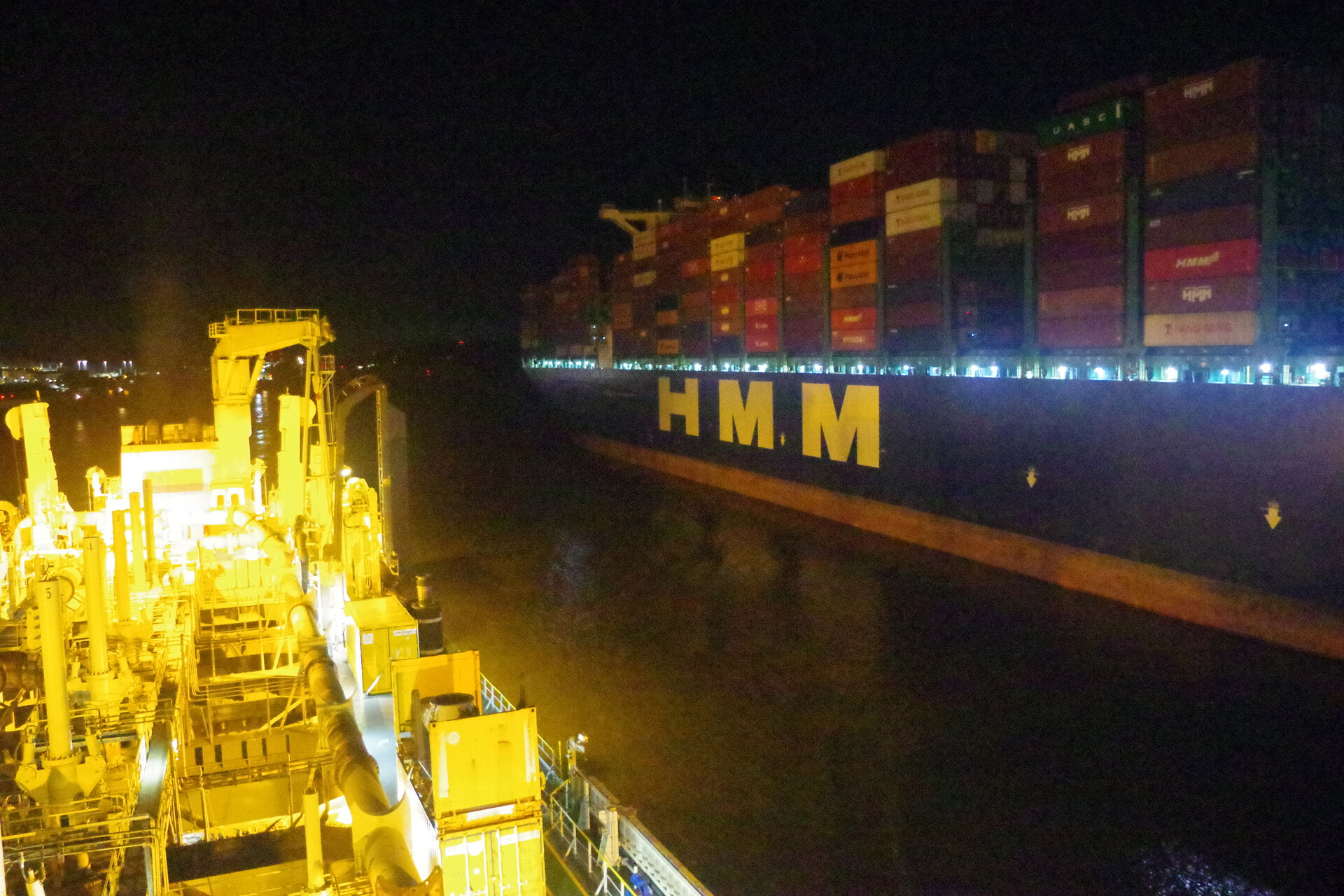 Bei Nacht aufgenommene Szene mit zwei großen Schiffen auf engem Wasserweg. Links im Bild ist das stark beleuchtete Baggerschiff KAISHUU mit zahlreichen technischen Aufbauten zu sehen. Rechts fährt ein riesiges Containerschiff mit der Aufschrift „HMM“ vorbei, beladen mit vielen Containern. 