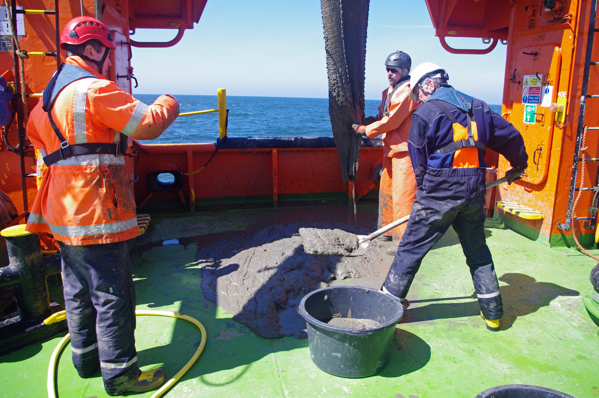 Drei Männer mit Schutzkleidung und Helmen stehen auf dem Deck eines Forschungsschiffs und entnehmen mit einer Schaufel eine nasse Sedimentprobe, die aus einem Netz auf den Boden gelangt. Im Hintergrund ist das offene Meer bei Helgoland zu sehen.