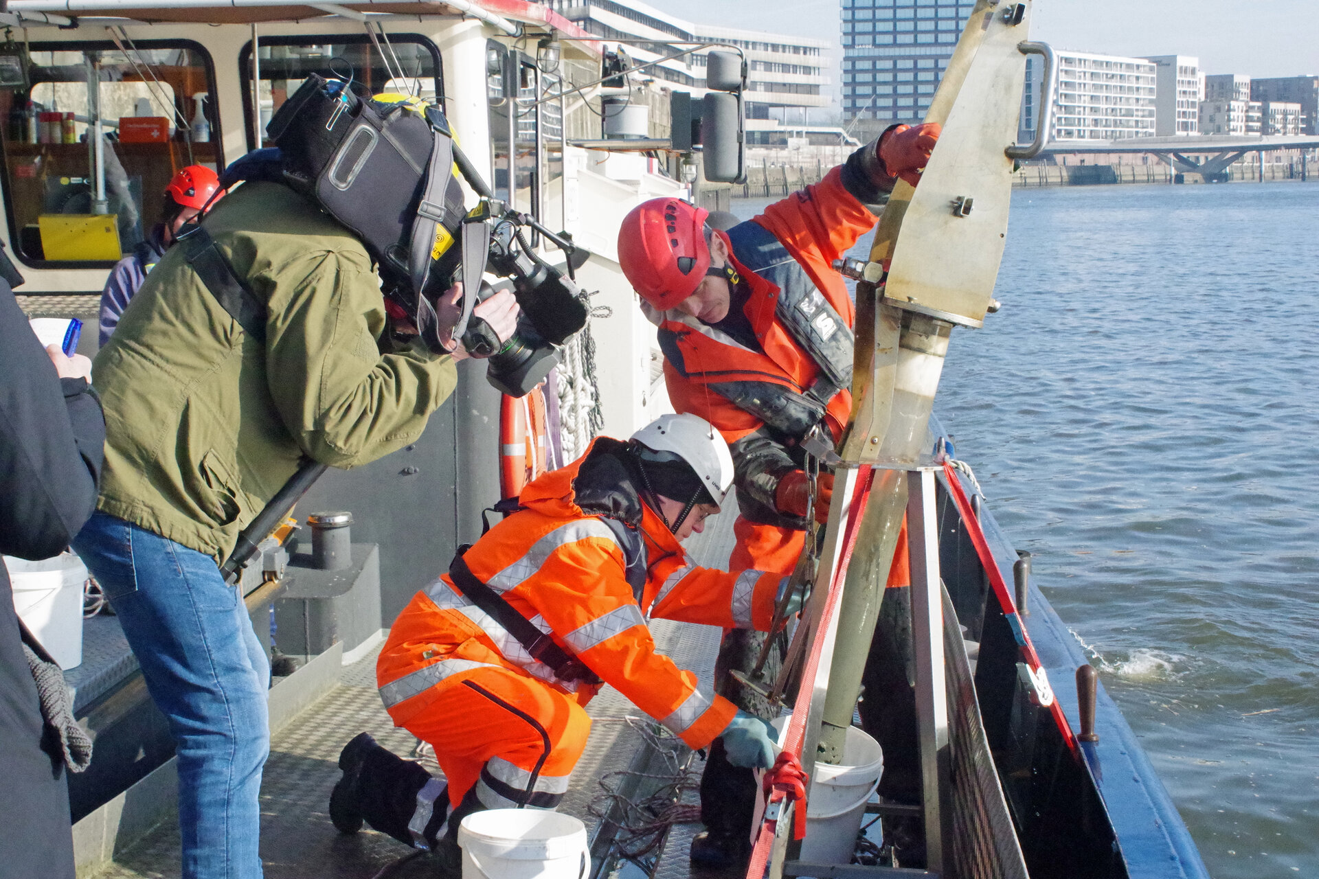 Zwei Personen auf einem Arbeitsschiff entnehmen eine Sedimentprobe aus der Elbe. Sie tragen orangefarbene Schutzkleidung und Helme. Eine dritte Person filmt die Szene mit einer professionellen Kamera. Im Hintergrund sind moderne Gebäude und eine Kaimauer des Hamburger Hafens zu sehen.